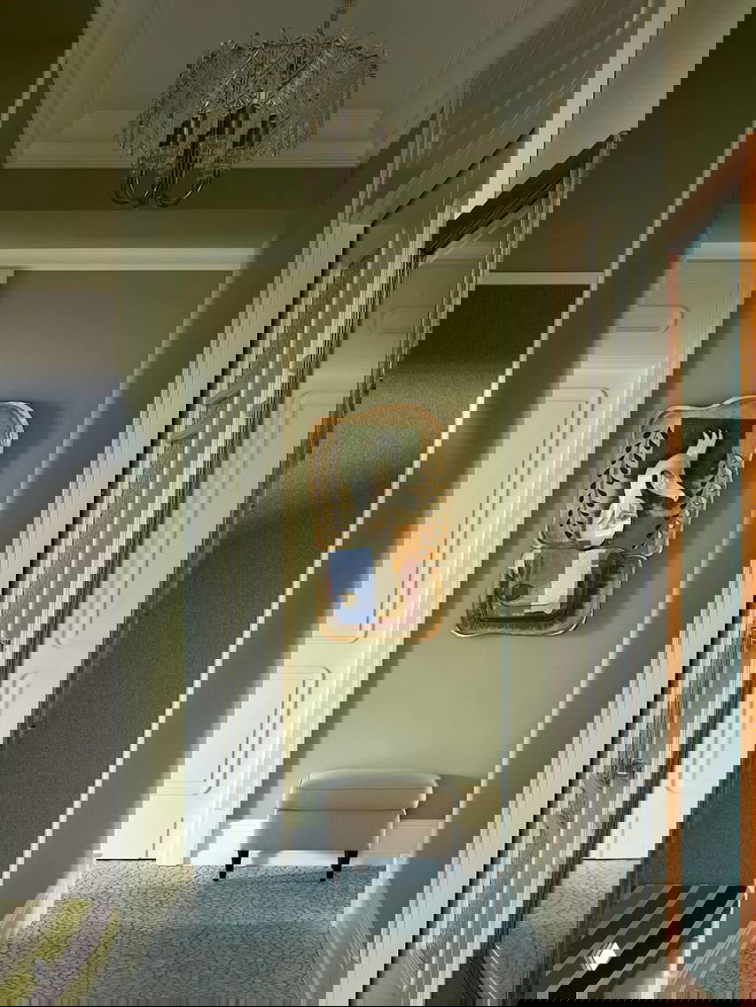 Узкий коридор, окрашенный в мягкий оливковый оттенок; небольшой пуф у стены, декоративное панно и хрустальная люстра над проходом.