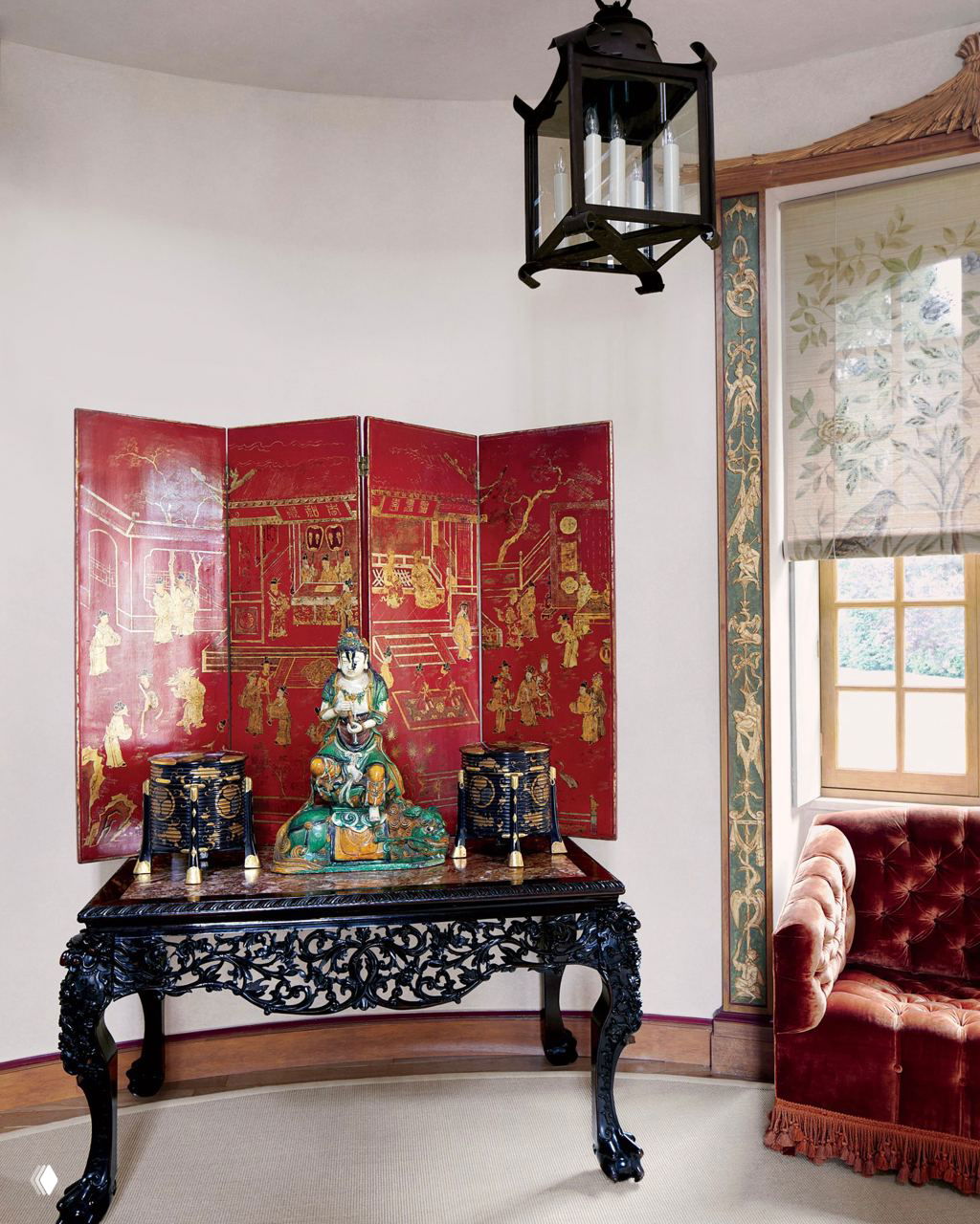 Уголок интерьера с резным консольным столом, красной ширмой в китайском стиле, фарфоровыми статуэтками и бархатным креслом у окна.