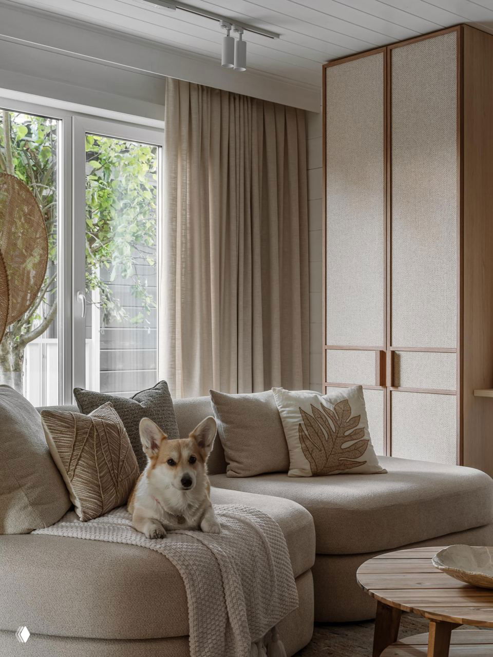 Гостиная в нейтральных тонах с бежевым диваном, лежащим корги на пледе и видом на озеро через большое окно — натуральные текстуры и мягкий свет.