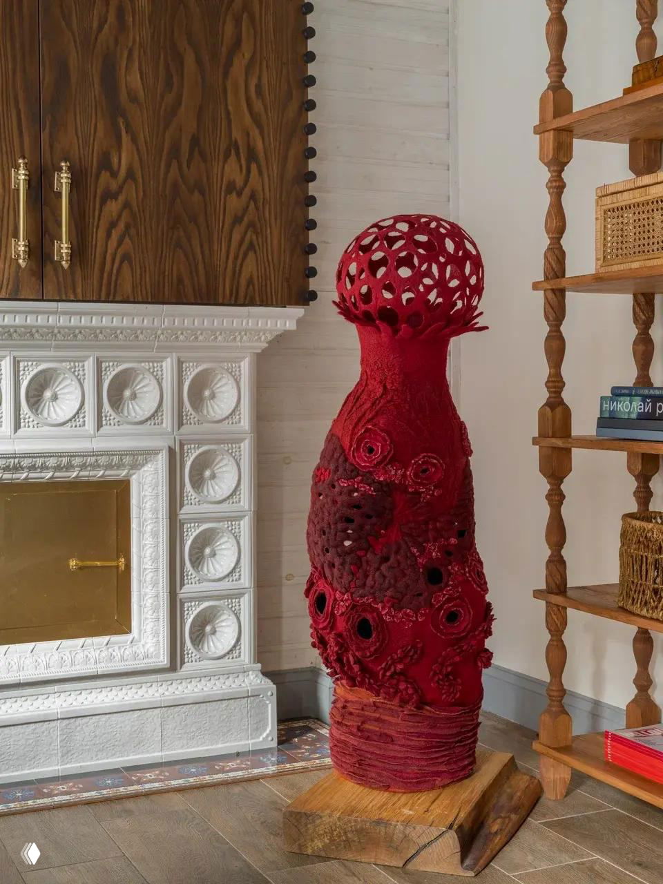 Высокая красная войлочная ваза-скульптура с множеством прорезей и объемной рельефной поверхностью стоит у классической печи, контрастируя с интерьером.