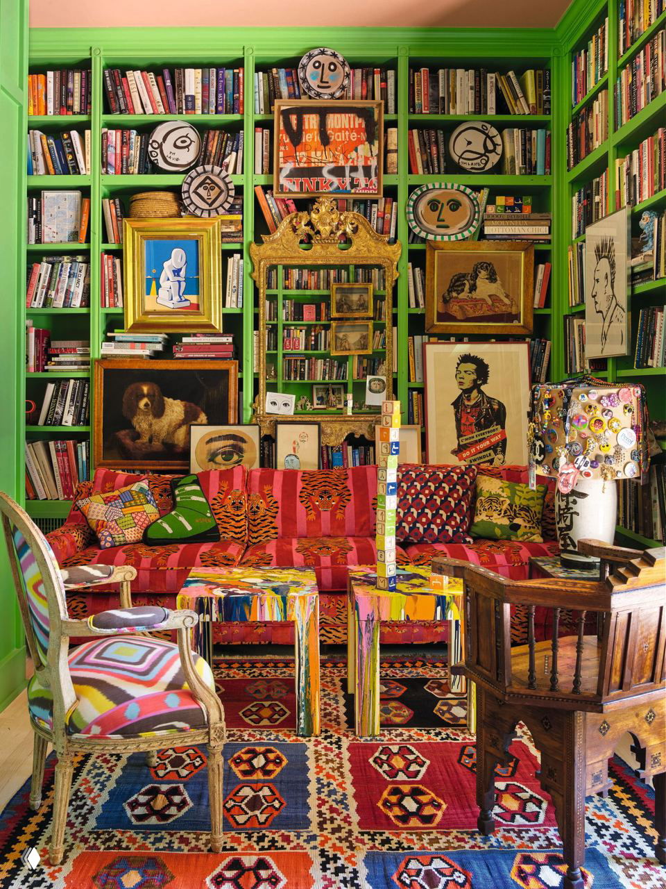 Зеленая библиотека с заполненными полками, золотым зеркалом и диваном в полоску на фоне яркого ковра и эклектичной коллекции картин и декора.