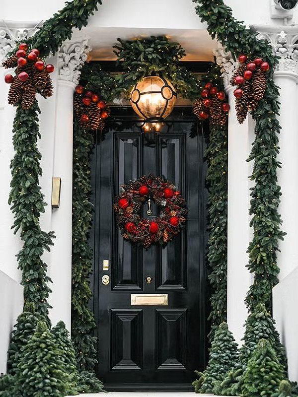Черная входная дверь украшена еловым венком с красными шарами и шишками, гирлянда обрамляет проём, ночное уличное освещение создаёт праздничную атмосферу.