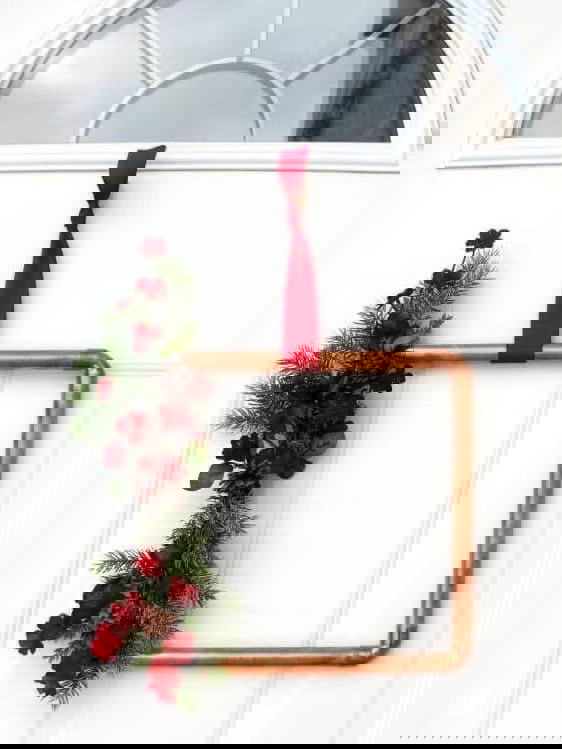 Дверная идея: медная рамка с привязанной красной лентой и веткой ели с ягодами и несколькими шишками в качестве минималистичного праздничного декора.