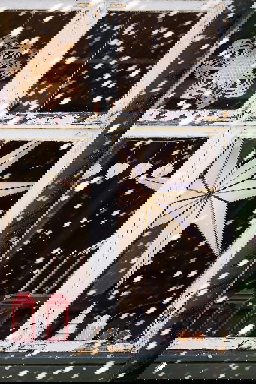 Новогодняя витрина за старым белым окном: бумажная звезда и тонированные свечи на фоне падающего снега и тёплого интерьера