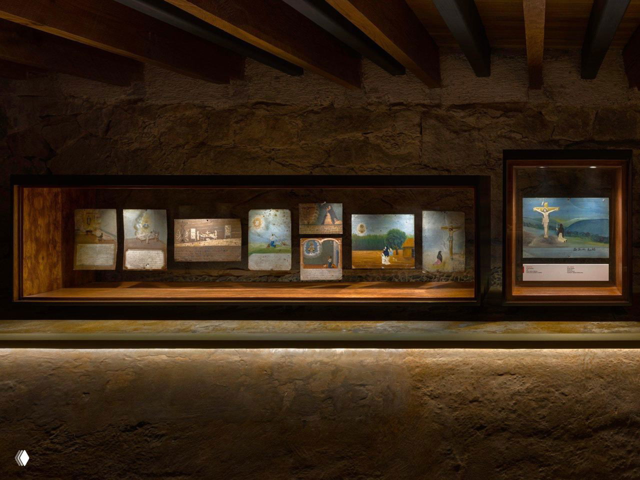 Тёмная музейная витрина с экспонатами и религиозными изображениями (ex-votos) в стеклянных рамках, подсветка подчёркивает детали экспозиции на каменной стене.