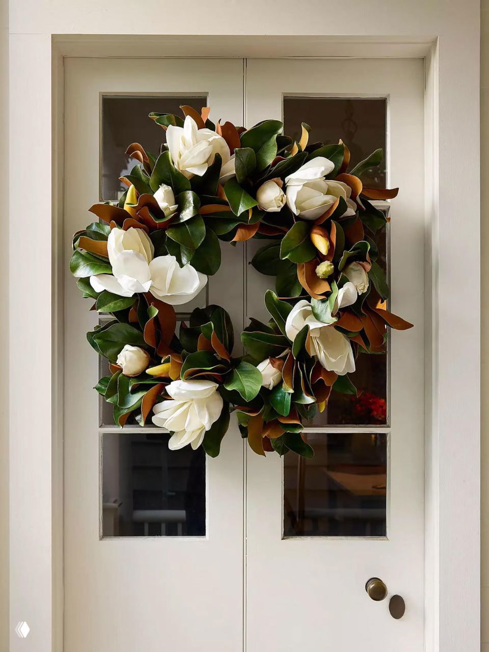Светлая дверь с крупным зелёным венком из листьев магнолии и белыми цветами, натуральный элегантный сезонный декор для минималистичного интерьера прихожей.
