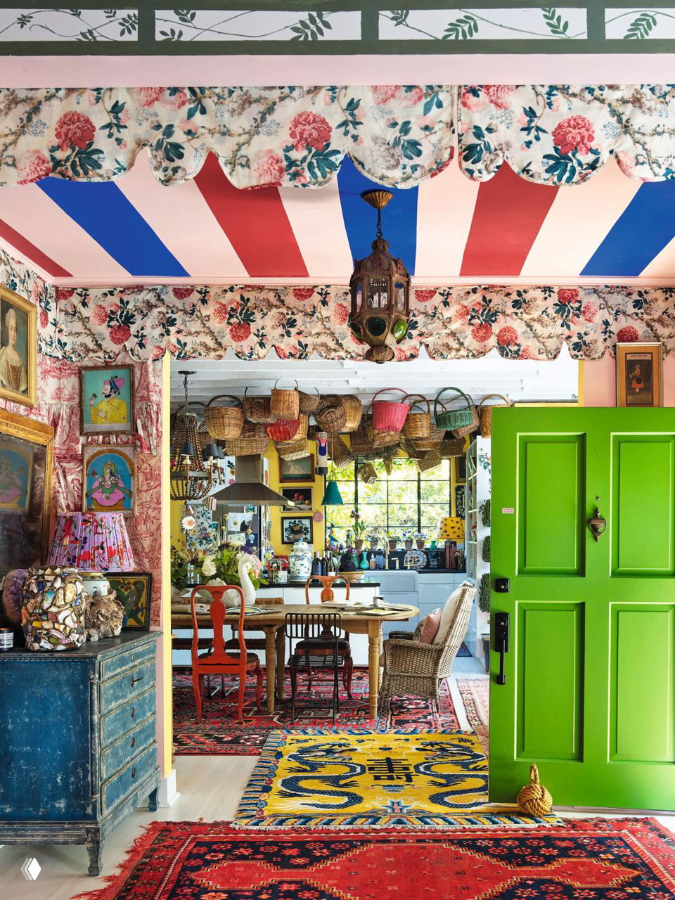 Вид через столовую на кухню: зелёная дверь, подвесные корзины над обеденным столом, яркие ковры и эклектичный текстиль в проходе между комнатами.