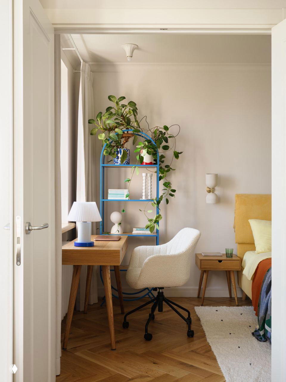 Рабочий уголок и часть спальни: белое кресло, деревянный стол, синяя стеллажная полка и фрагмент жёлтой кровати с ярким покрывалом.