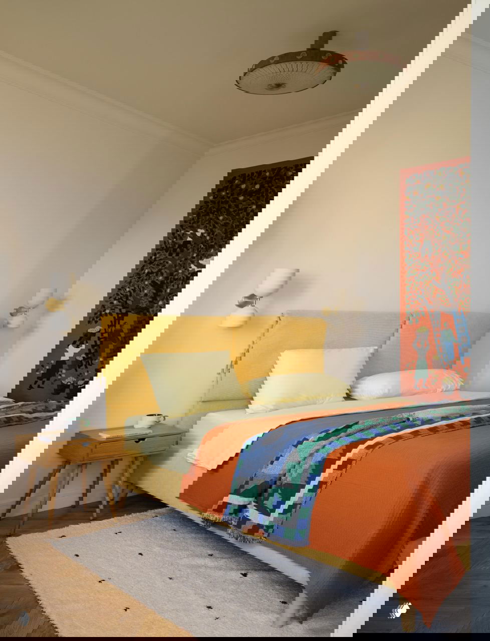 Спальня с жёлтой обивкой кровати, оранжевым покрывалом, декоративным текстилем и художественным панно на стене; тёплый паркетный пол.