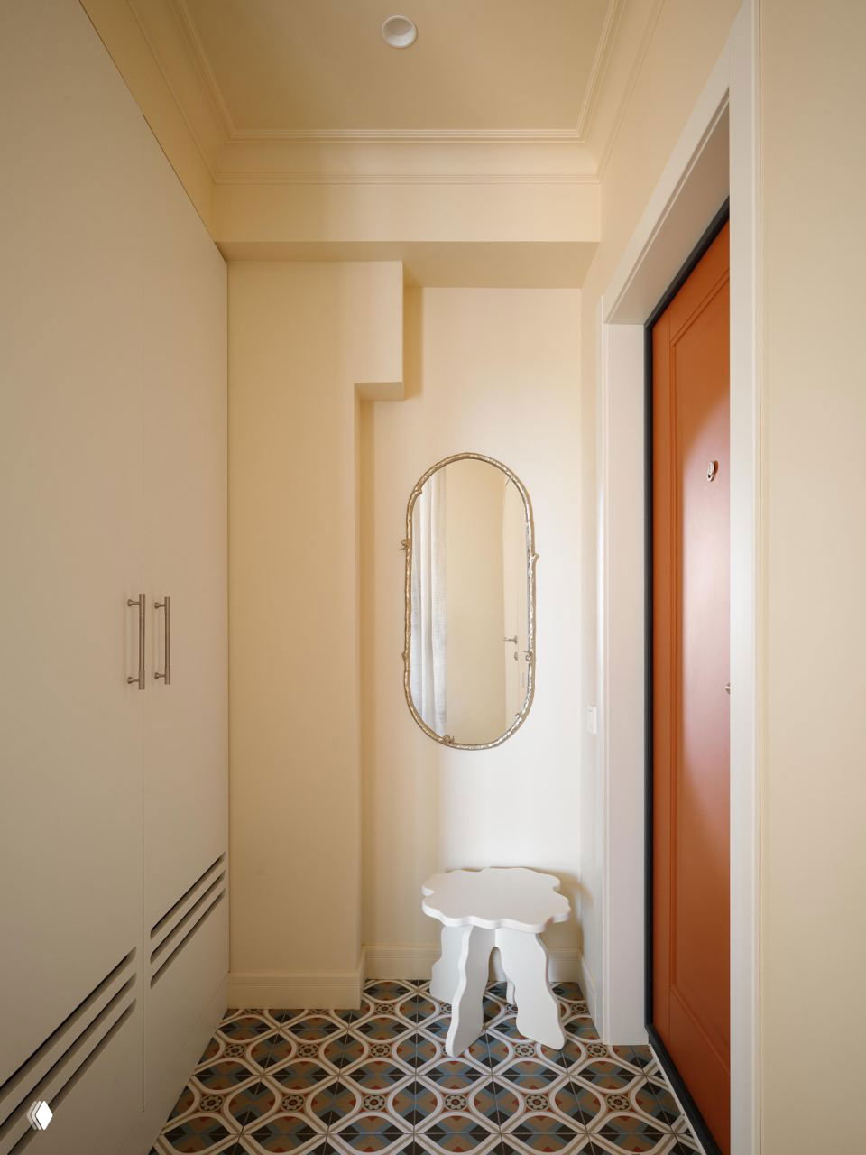 Прихожая с узким белым табуретом, овальным настенным зеркалом и входной дверью терракотового цвета; узорчатая плитка на полу создаёт акцент.
