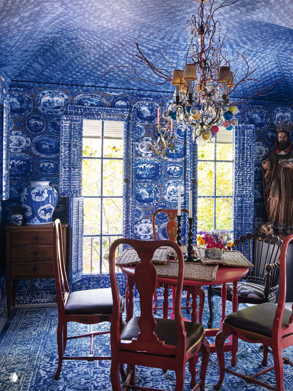 Синяя столовая полностью покрыта сине‑белым узорчатым текстилем и фарфором, с красными деревянными стульями и декоративной люстрой над столом.