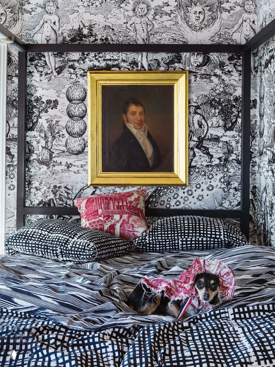 Спальня с черно‑белым принтом на стенах и текстиле, четырехстоечная кровать, старинная картина в золотой раме и собака, устроившаяся на подушках.