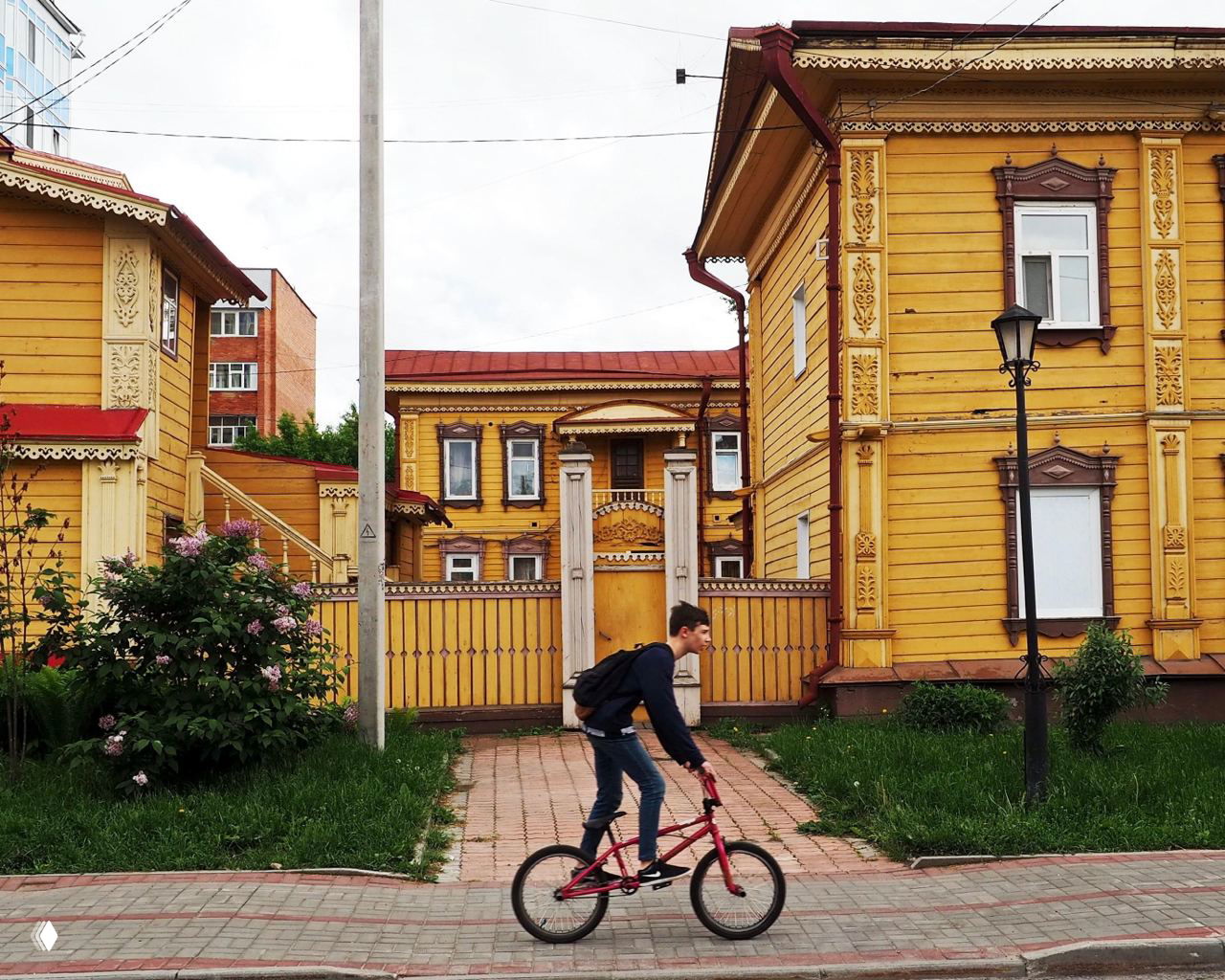 Улица Томска с крупным жёлтым деревянным особняком и резными деталями фасада; внизу кадра велосипедист едет по тротуару в городской среде.