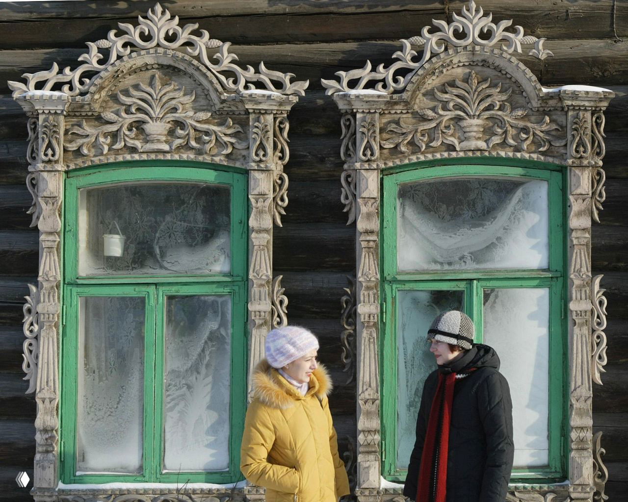 Два человека разговаривают у старого деревянного дома с зелёными рамами окон и богато резными наличниками — фрагмент традиционного томского зодчества.