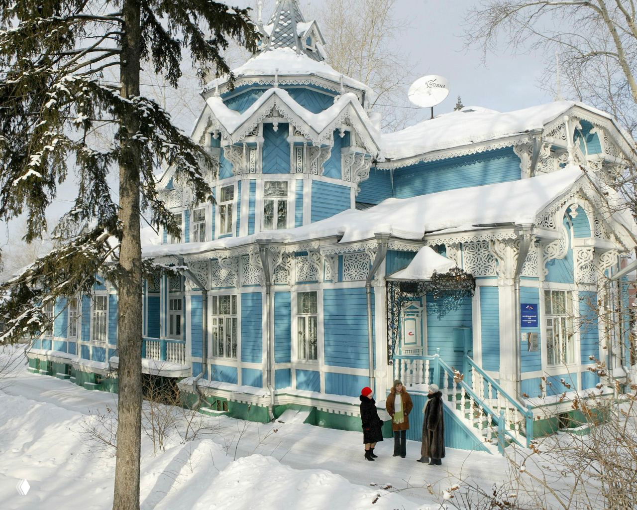 Сине-белый деревянный особняк Томска с изящной резьбой по карнизам и верандой, окружённый сугробами и людьми у входа в зимний день.