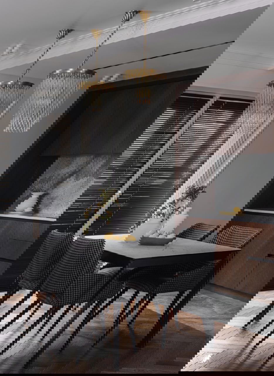 Обеденная зона с мраморным столом и шестью стульями в тёмно-серой фактуре, над столом висят две крупные хрустальные люстры, вид на кухонные фасады.