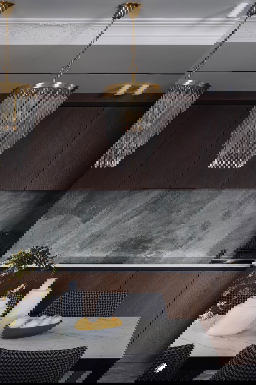 Кухонный фартук из серого камня, тёмные деревянные шкафы до потолка и декоративные подвесные люстры со стеклянными трубками вблизи рабочей зоны.