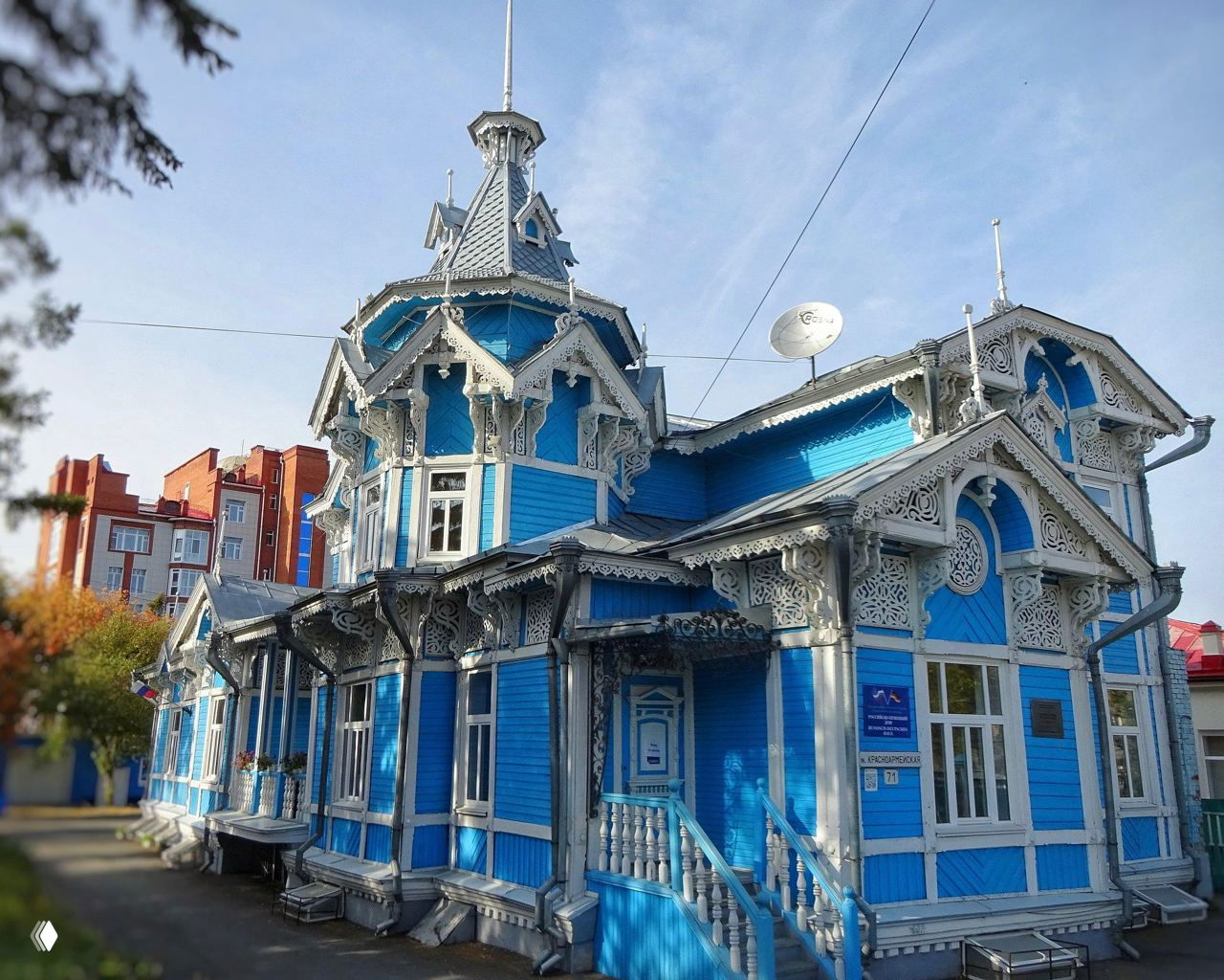 Колоритный голубой деревянный дом с резными украшениями и башенкой на переднем плане; фасад демонстрирует богатую орнаментальную резьбу по дереву.