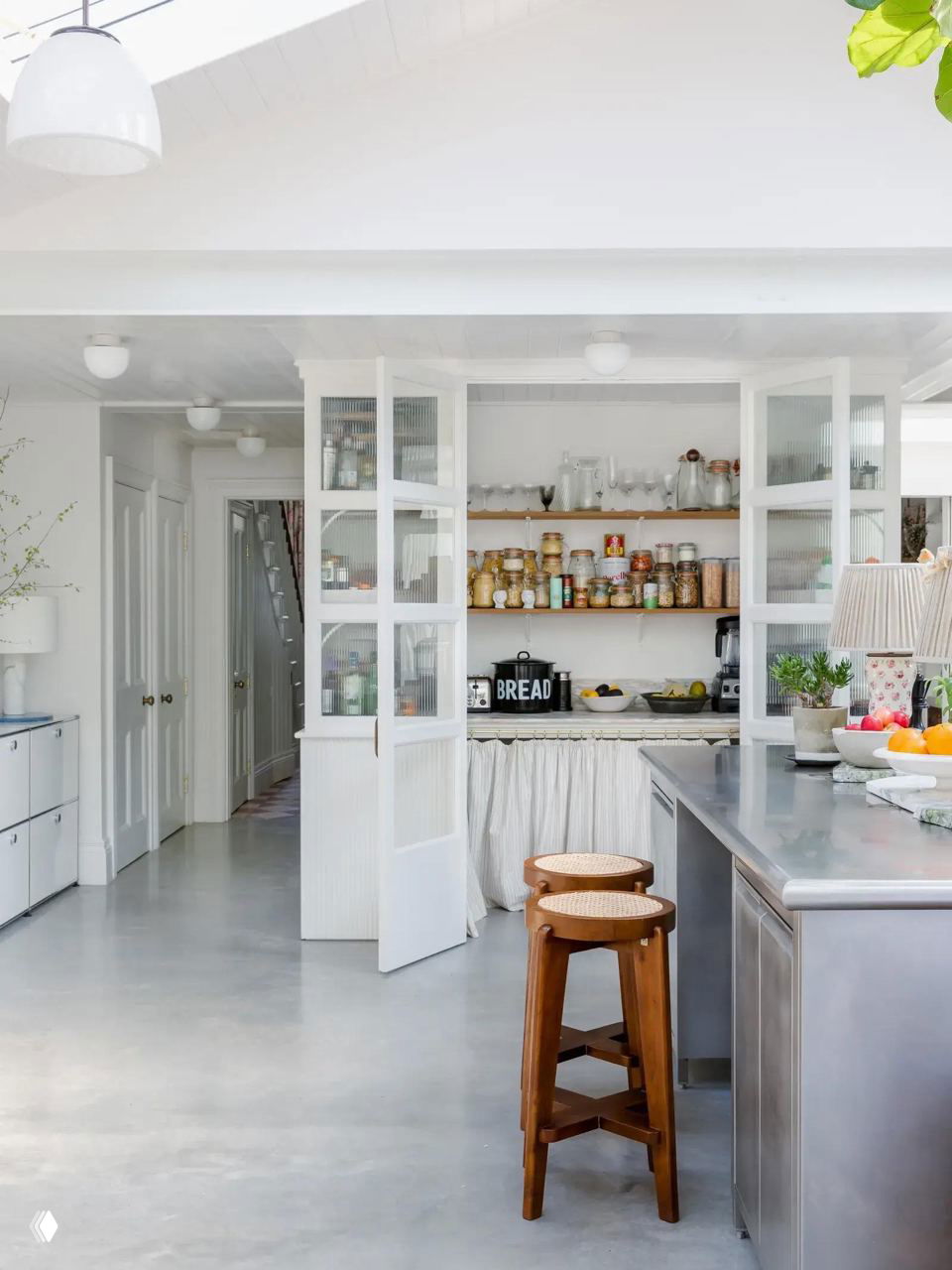 Светлая кухня с низким островом и стеклянными дверцами кладовой, где видны аккуратно расставленные банки и контейнеры — идея для организованного хранения.