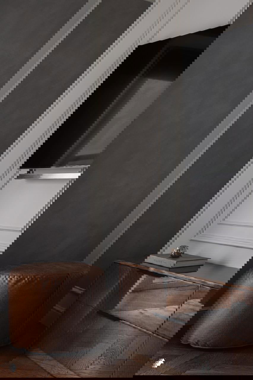 Интерьерная деталь: коричневый круглый пуфик, глянцевый комод и телевизор на классической стеновой панели с молдингом и лепниной.