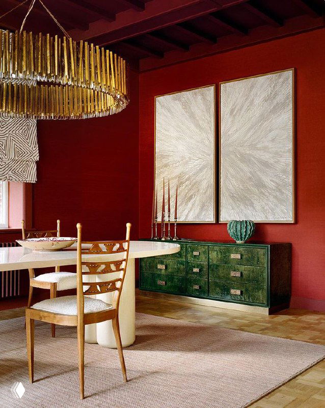 Столовая с насыщенными бордовыми стенами и кессонным потолком, деревянный стол и стулья на светлом ковре; зелёный комод и золотая люстра создают яркий интерьер.