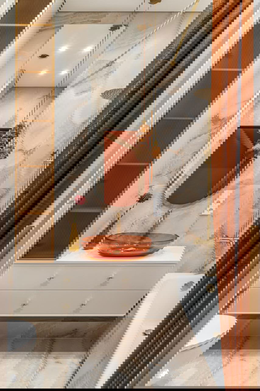 Санузел с мраморной облицовкой, белой тумбой и терракотовой стеклянной раковиной; золотые смесители и занавеска в тёплых оттенках.