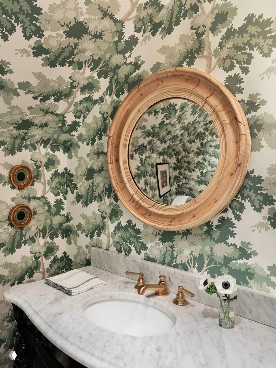 Обои с узором ветвей и листьев в зелёной палитре, круглое деревянное зеркало и мраморная столешница с встроенной раковиной.