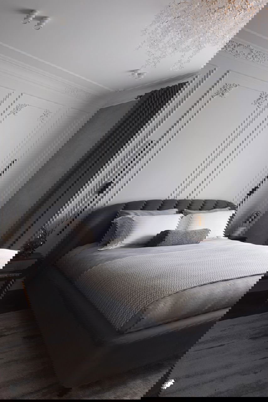 Главная спальня в спокойной серой гамме: большая кровать с мягким изголовьем, текстиль в тон и декоративная лепнина по периметру потолка и стен.