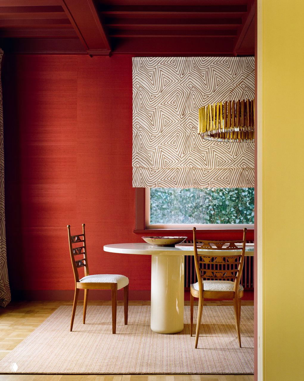 Кадр столовой с круглым белым столом у окна, бордовые стены и текстурные римские шторы на фоне; видна часть солнечной жёлтой стены рядом.