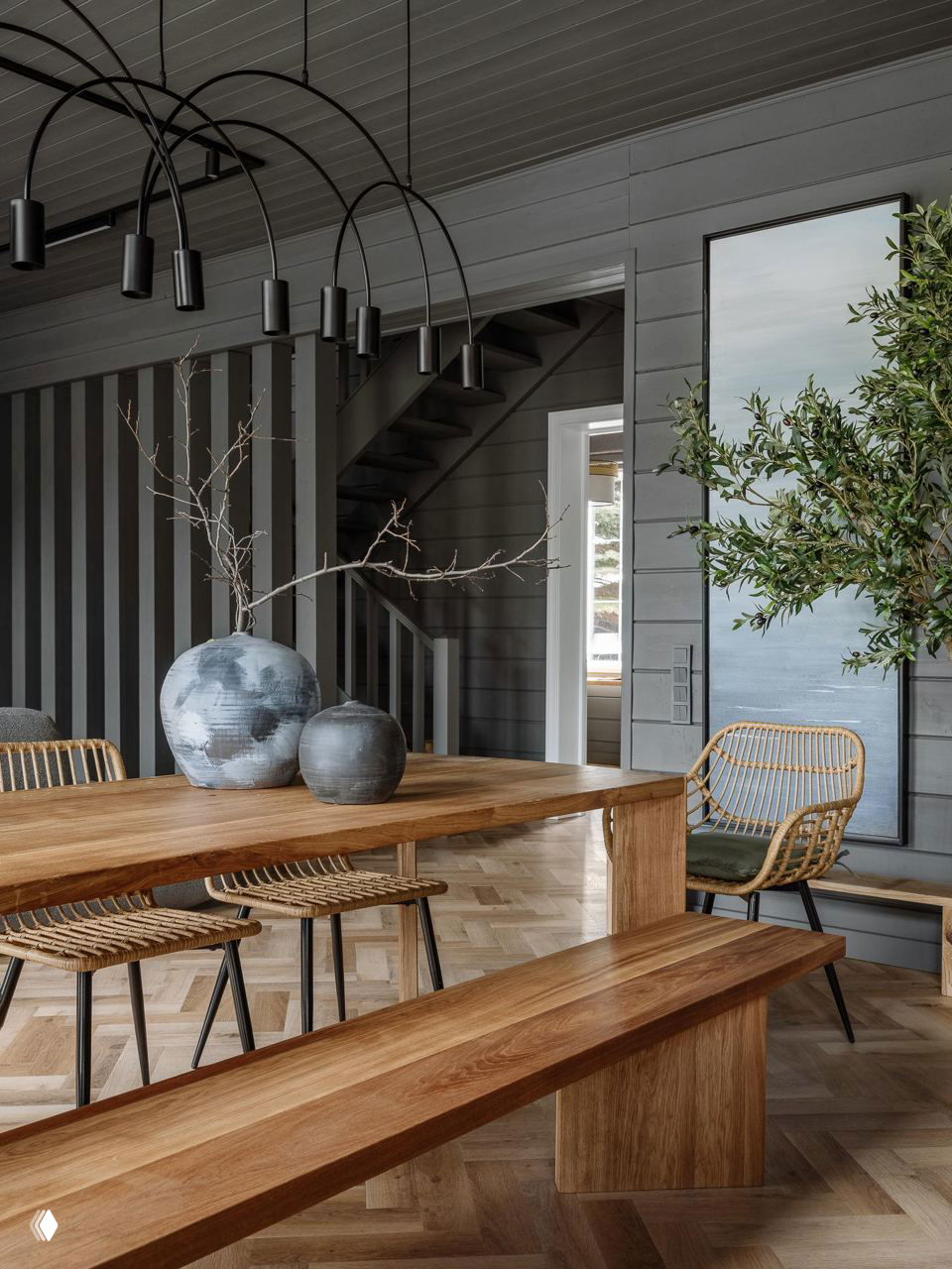 Столовая с большим деревянным столом и скамьями на фоне серых стен и лестницы; натуральное дерево и нейтральные тона формируют спокойную нейтральную палитру.