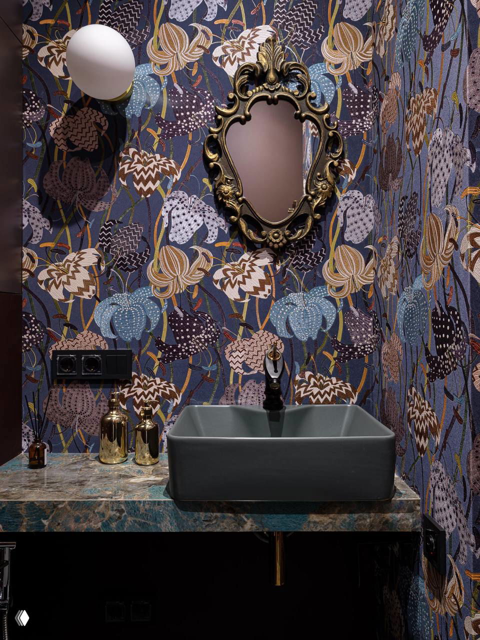 Обои с флористическим узором в холодной гамме, декоративное золочёное зеркало и тёмная прямоугольная раковина на фоне.