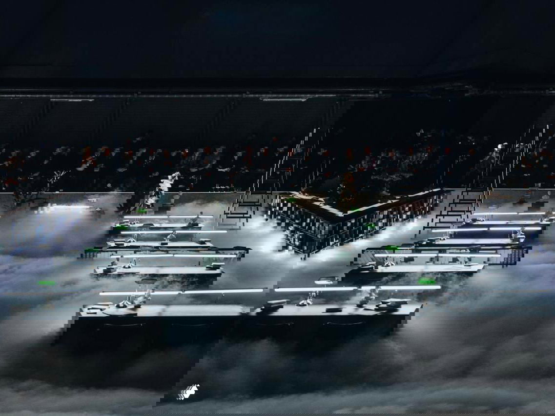 Широкий ракурс оперной инсталляции в читальном зале: ряды столов с зелёными лампами, плотный театральный туман и зрители по периметру сцены.