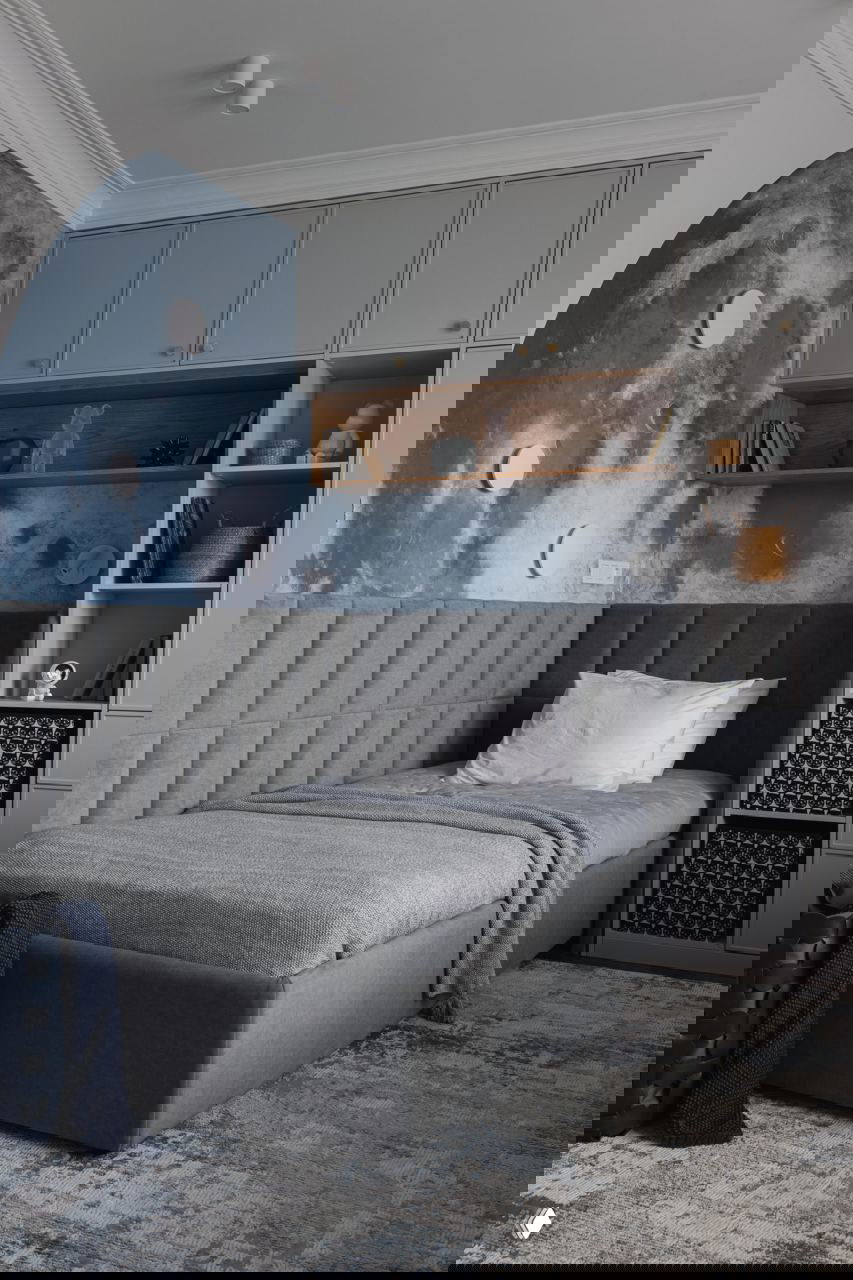 Детская комната с настенной росписью в виде луны, встроенными шкафами над кроватью, мягким серым изголовьем и функциональными полками для хранения.