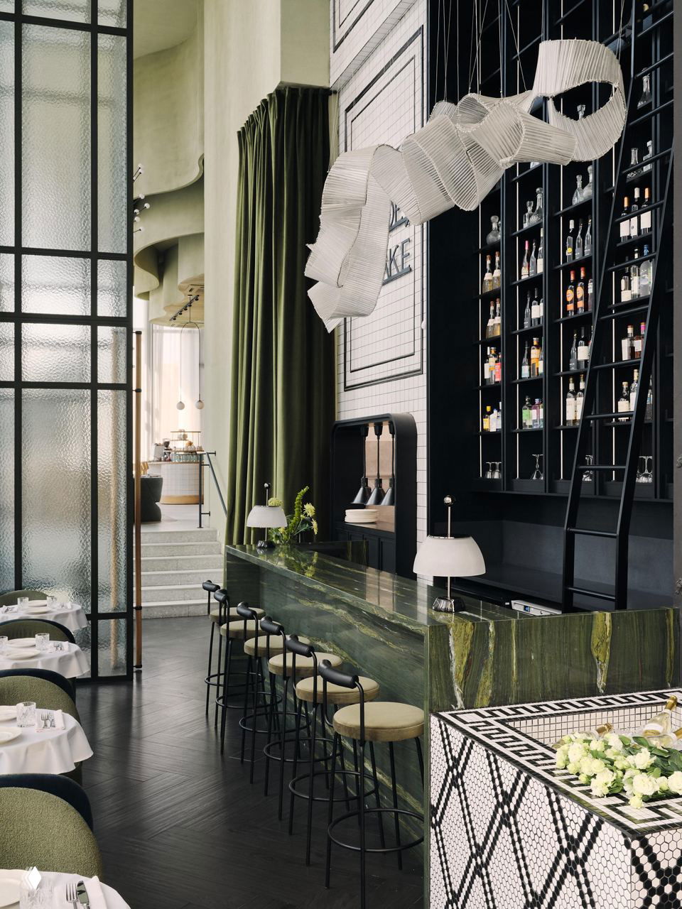 Барная стойка из зелёного камня, высокий бар и лестница к полкам с бутылками, в композиции видны крупные шторы и элементы перегородки.
