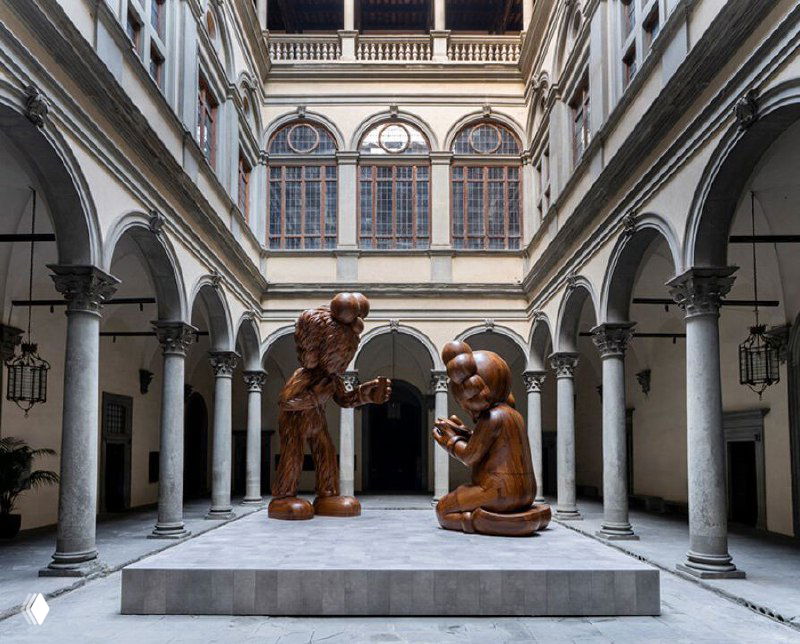 Крупный план композиции «Послание» во дворе: стоящий персонаж Companion и сидящая BFF из дерева на фоне архитектуры палаццо Строцци.