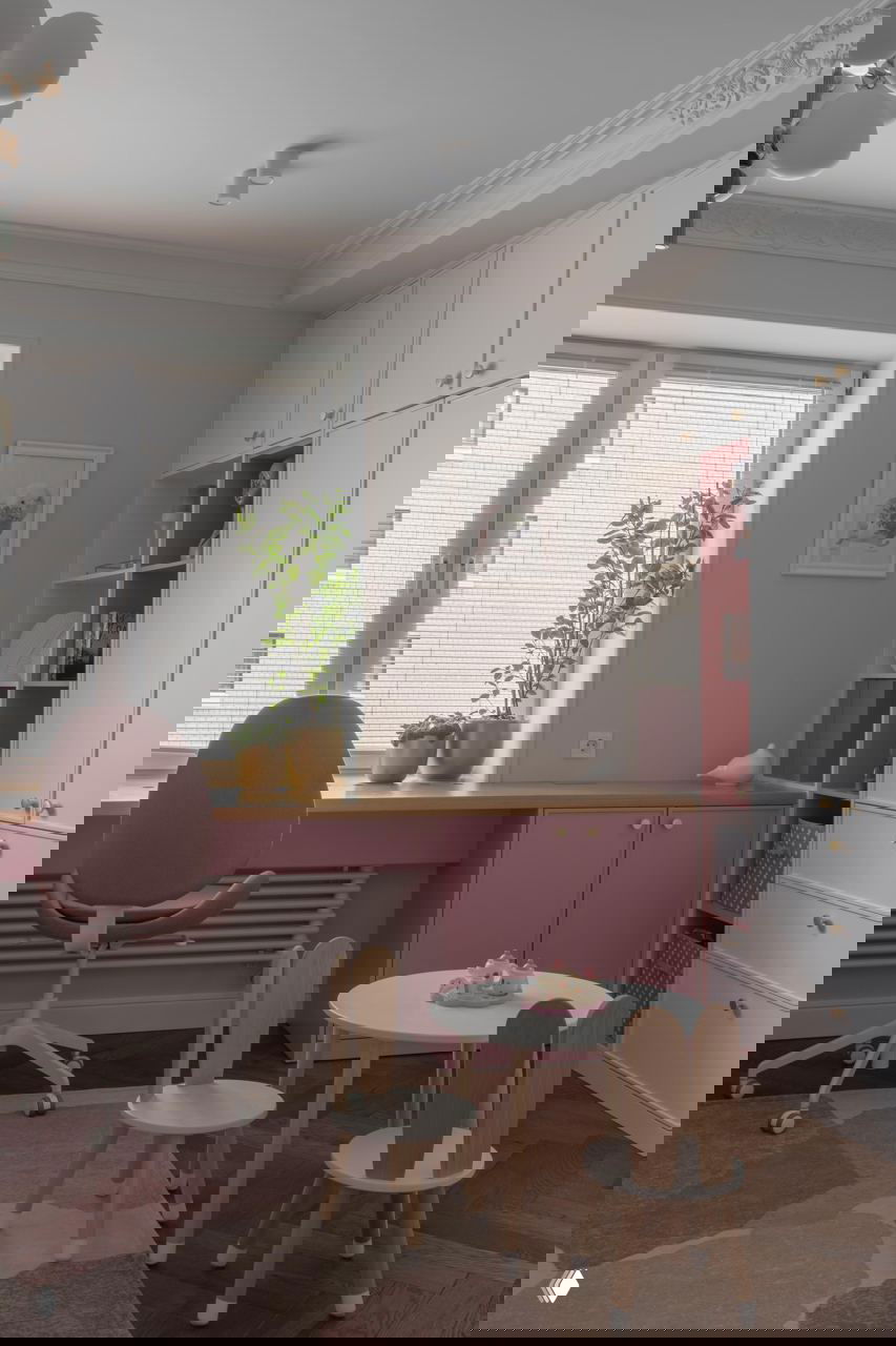 Рабочая зона у окна с подоконником-столом, розово-серые шкафы и детский стол с мини-стульями — светлая функциональная ниша для учебы и творчества.