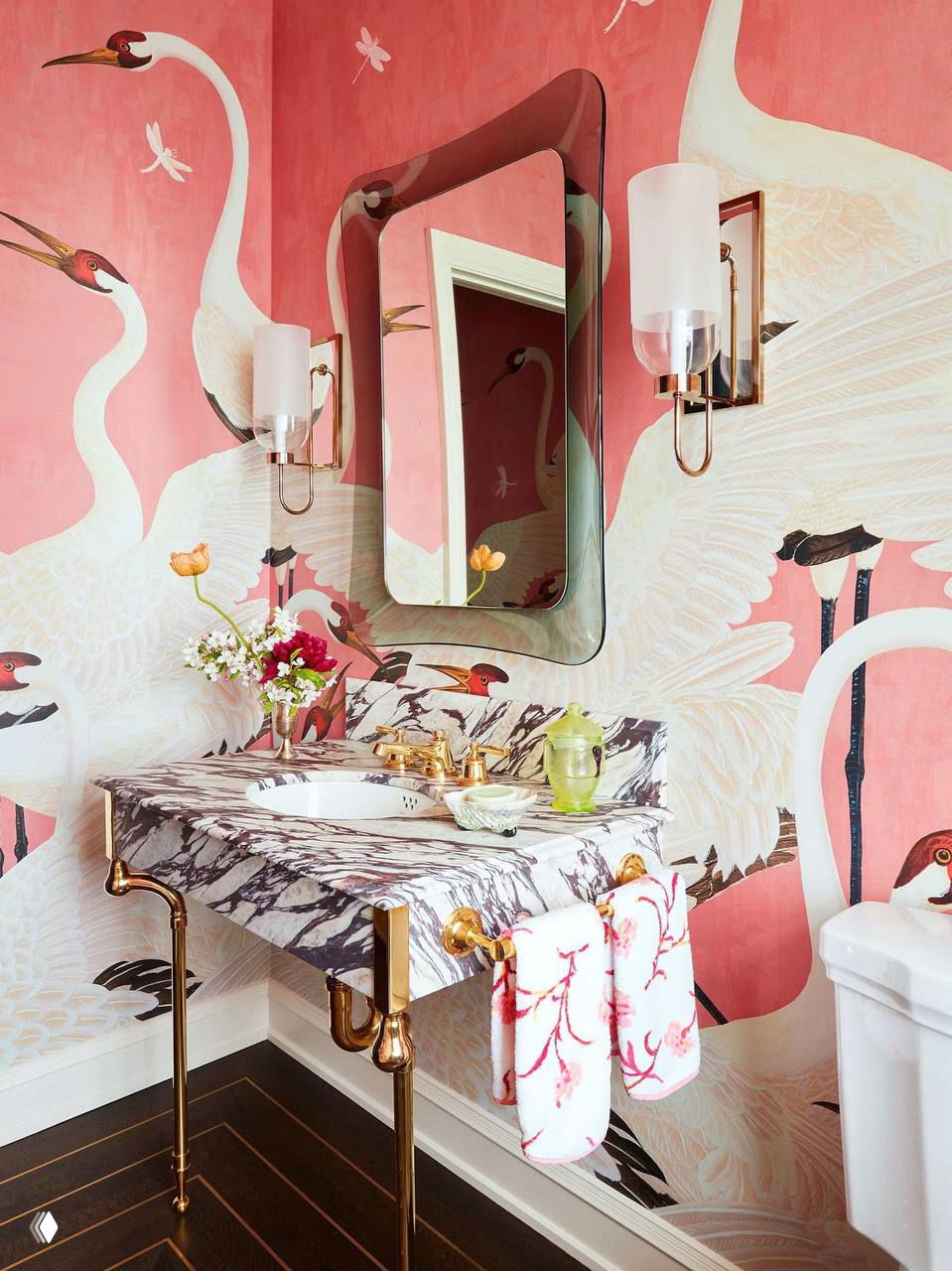 Розовые обои с изображением журавлей и растительными мотивами, мраморная столешница, золочёные смесители и декоративные полотенца.