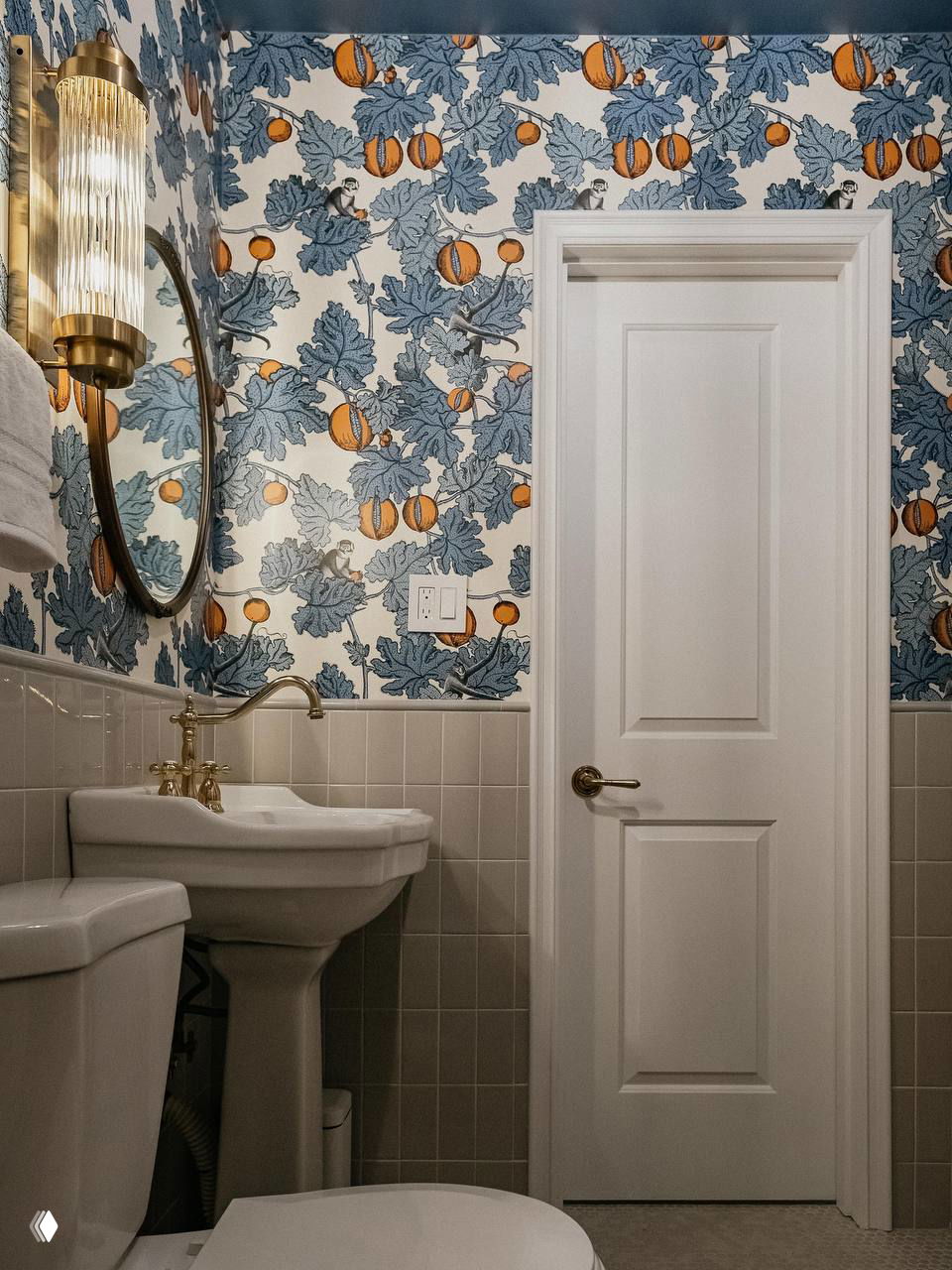Обои с рисунком листьев и тыкв в голубой и оранжевой гамме, видна высокая белая пьедестальная раковина и настенный круглый светильник.