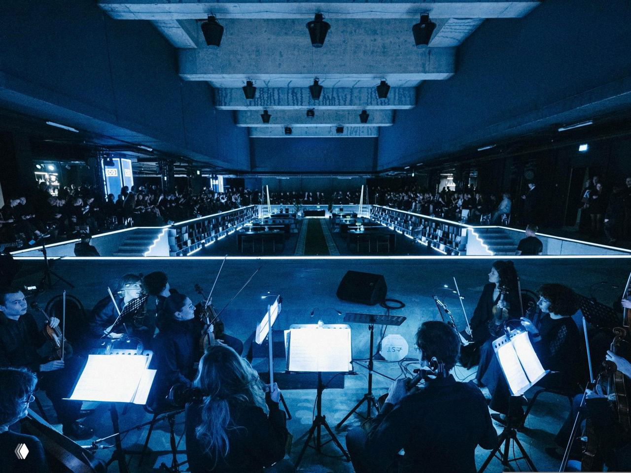 Вид с оркестровой ямы: музыканты на фоне инсталляции, обрамлённой полками и подсветкой, зрители размещены вокруг сценического пространства.