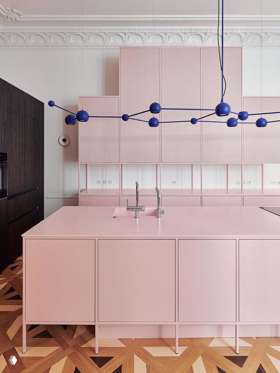Розовая кухня с широким островом, синие сферические подвесы, декоративная лепнина на стенах и геометрический паркет в интерьере квартиры.