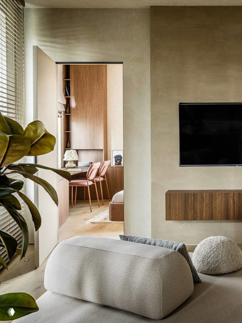 Вид из гостиной в прилегающую рабочую или столовую зону: мягкие формы дивана, деревянные шкафы и натуральная отделка стен и пола в тёплых оттенках.