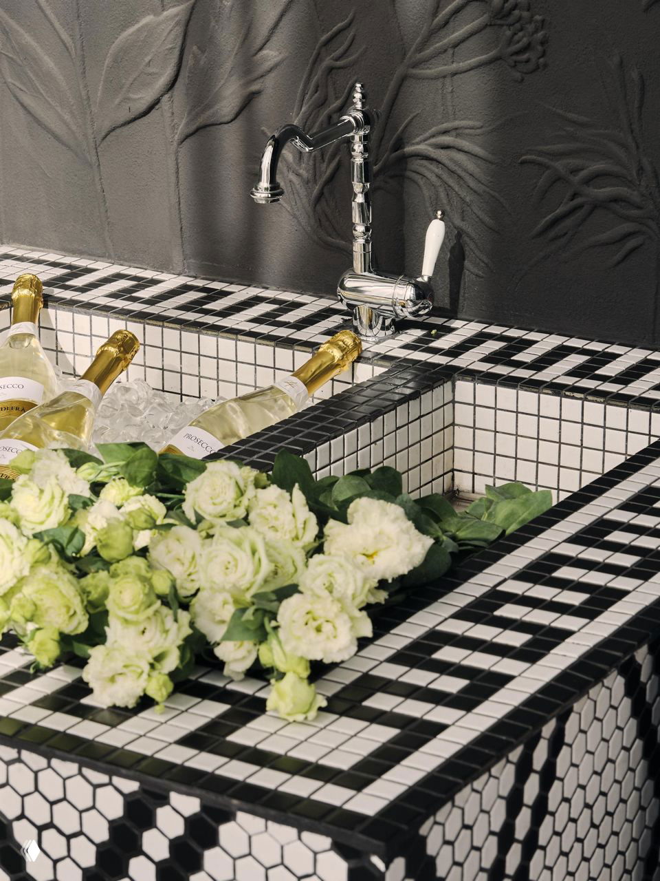 Мозаичная чаша-ледник для шампанского в чёрно-белой плитке, наполненная бутылками и украшенная цветами, деталь бара ресторана.