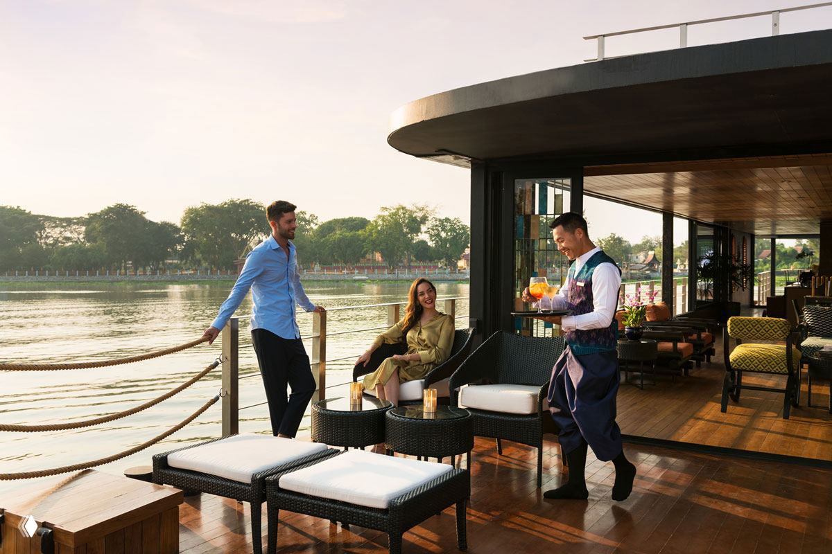 Открытая палуба баржи с креслами и столиками: официант подаёт напитки, солнечные блики на деревянной палубе и спокойная река Чхаопрайя на заднем плане.