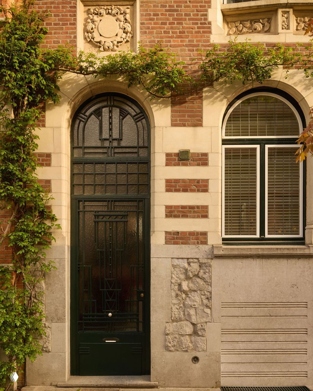 Фасад исторического таунхауса в Кортрейке: арочная чёрная дверь, кирпичная кладка, каменные декоративные детали и вьющиеся растения вокруг входа.