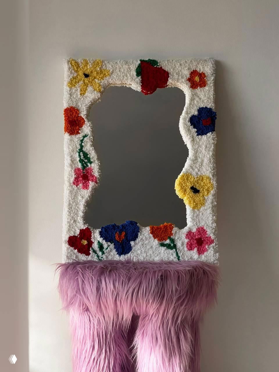 Зеркало в рамке с яркими цветочными тафтинг‑фрагментами и грубой текстурой, расположенное над консолью с розовым пушистым покрытием как интерьерный акцент.