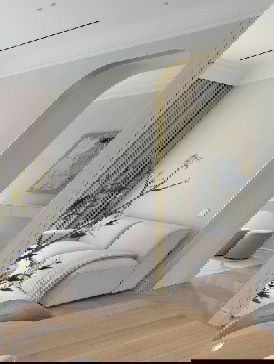 Угол гостиной с изящной аркой в переходе, бледно-бежевый диван и деревянный паркет ёлочкой, декоративные шторы и минималистичная планировка.