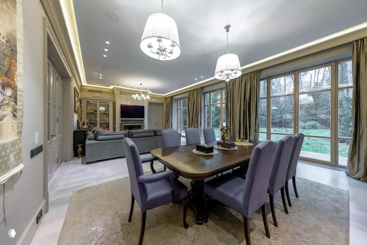 Широкий ракурс столовой и гостиной с крупным окном на сад, мягкой зоной для отдыха, люстрами и световой подсветкой по периметру потолка.