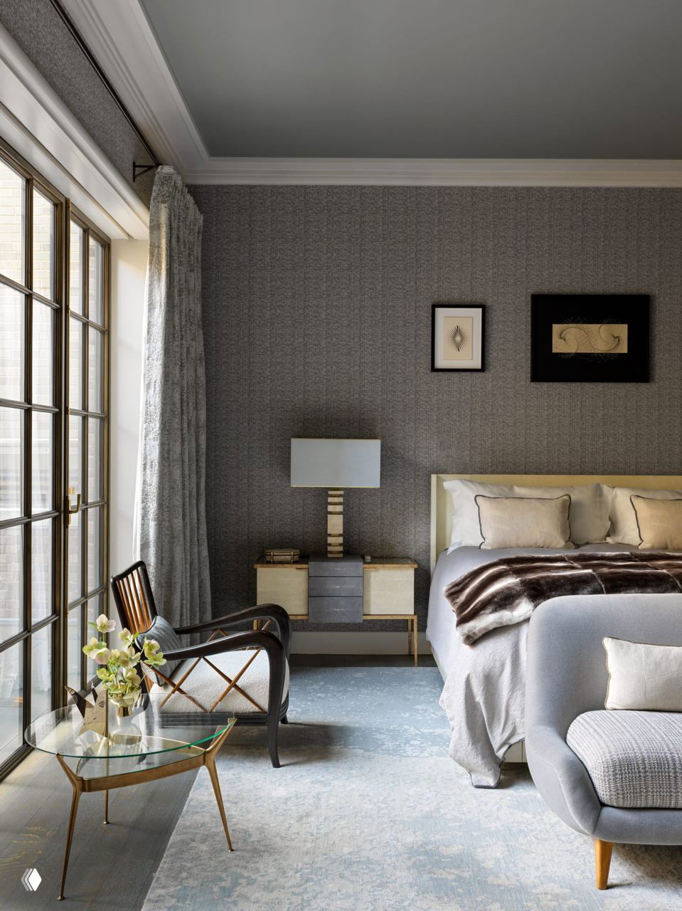 Спальня с мягкой кроватью, текстурными стенами и больших окон с бронзовыми рамами; кресло у круглого стеклянного столика.