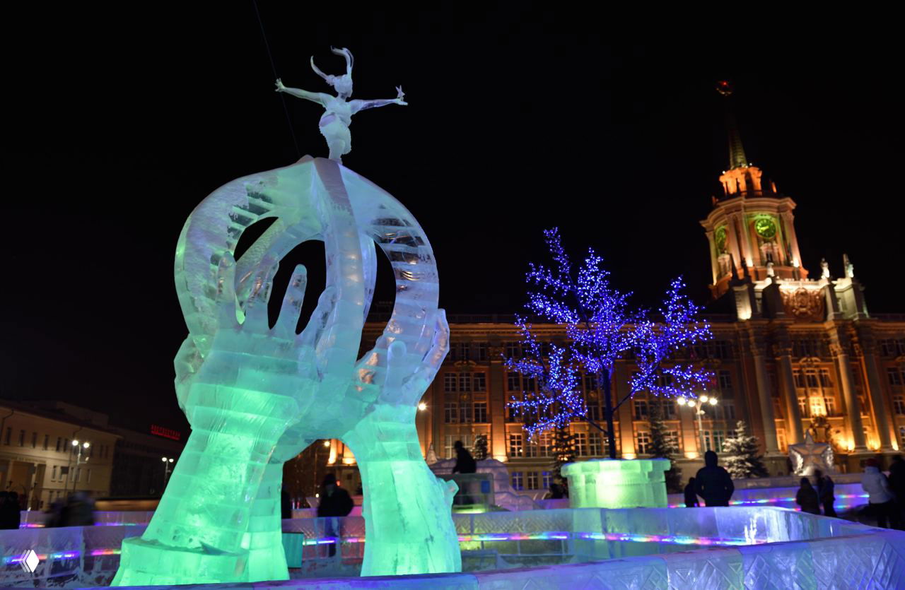 Подсвеченная ледяная скульптура на центральной площади Екатеринбурга на фоне ночных огней и архитектуры, праздничная зимняя атмосфера.
