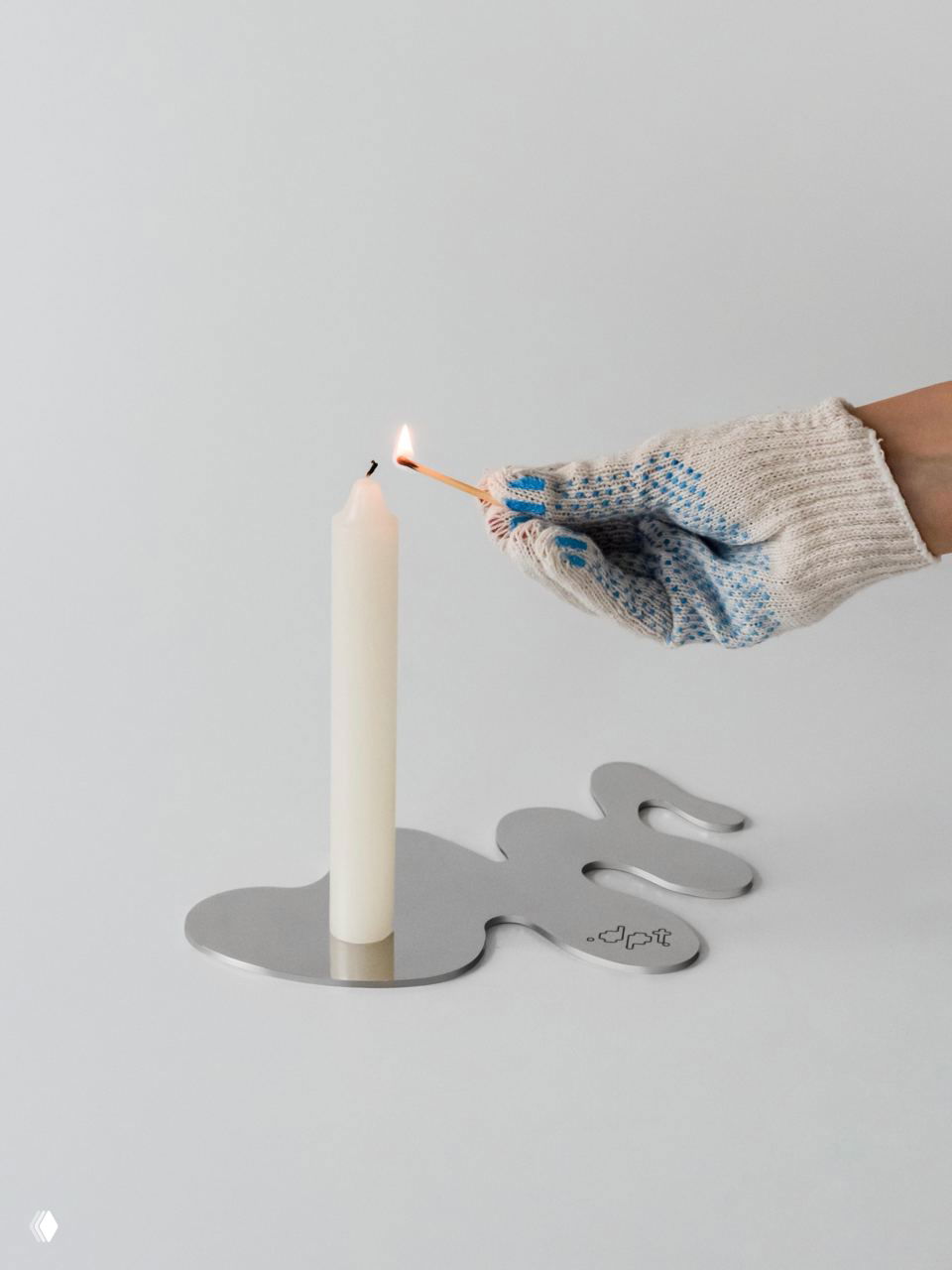 Рука в рабочей перчатке подносит зажженную спичку к высокой свече, установленной в металлическую подставку в форме «лужи», демонстрация контраста воска и металла.
